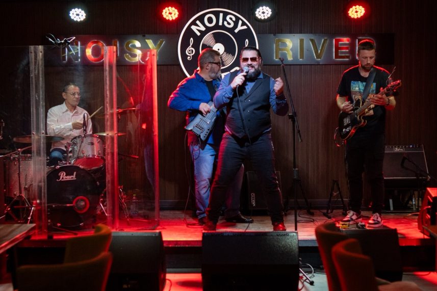 Энергичное выступление ZebraWood Blues Band в Noisy River — вокалист и музыканты на сцене под ярким клубным освещением с логотипом Noisy River.